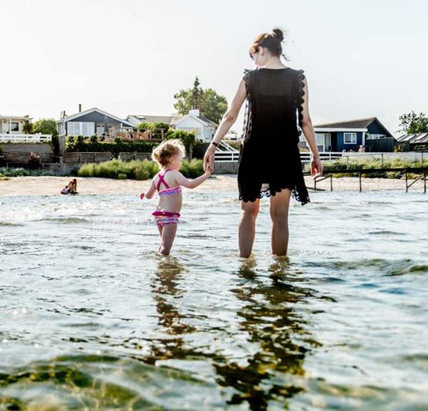 En mor går med en lille pige i hånden og sopper i vandet langs strandkanten.