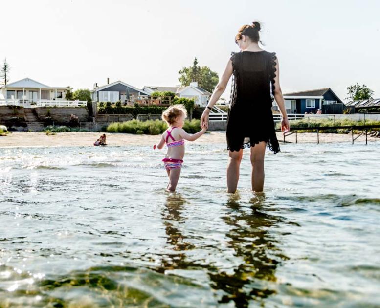 En mor går med en lille pige i hånden og sopper i vandet langs strandkanten.