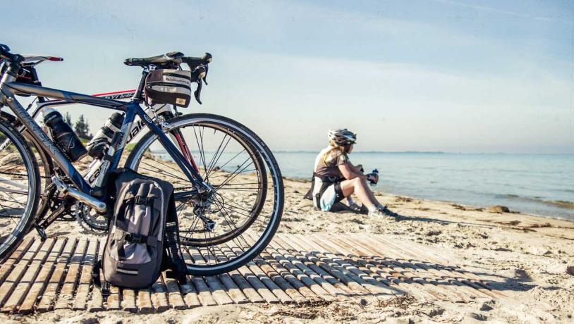 To parkerede cykler på strand. I baggrunden sidder de to cykelryttere i sandet i vandkanten og holder pause.