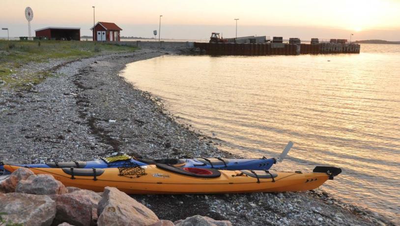 På en strandbred ligger to kajakker i forgrunden. I horisonten af billedet kan man se en  strandspids hvor der står et lille rødt hus foran solnedgangen. 