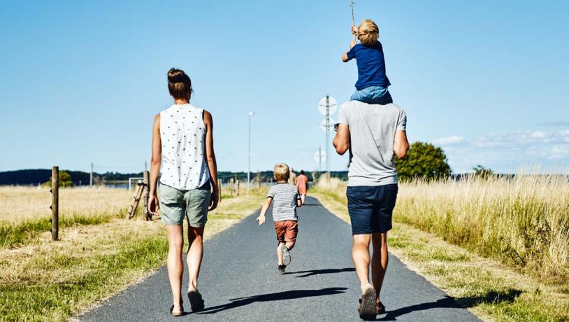 En familie på fire går ned ad en landevej på en solrig dag. Manden til højre bærer et lille barn på sine skuldre. Mellem dem går et andet barn. De er omgivet af åbne marker, og himlen er klar og blå.