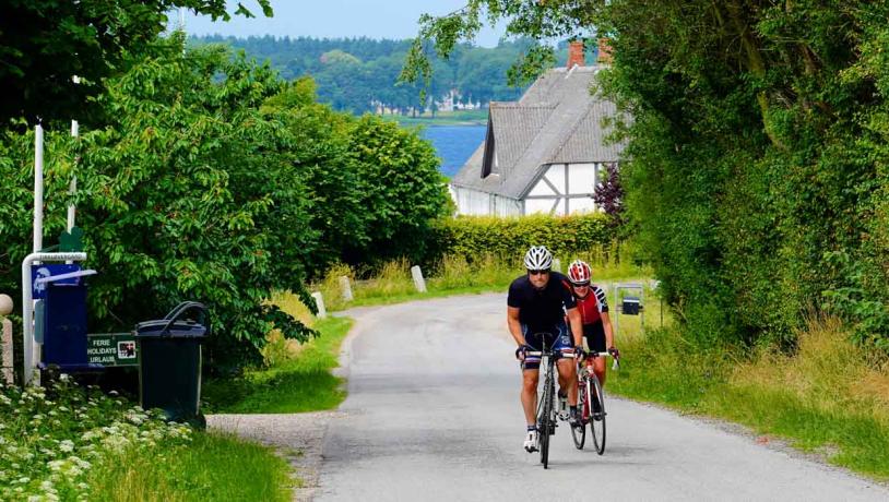 To cyklister kører af sted på asfaltvej med grønt til alle sider, hvidt bindingsværkshus og hav i baggrundet.