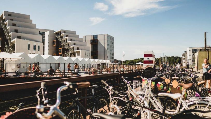 Havnebadet i Odense på en solskinsdag med blå himmel. Der er badende mennesker i havnebadet og sommerklædte mennesker, som går ude foran. Der står en masse cykler i forgrunden.