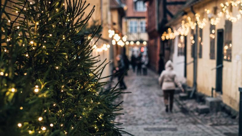 I højre side står et juletræ med julelys. Bag juletræet ses en sløret brostensgade der går ned mellem to rækker af gamle bindingsværkshuse. På husene hænger julelys, og på gaden går et par mennesker.