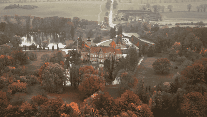 Dronefoto af et slot med spir og tårne placeret midt i et efterårsfarvet landskab i grønne og orange farver