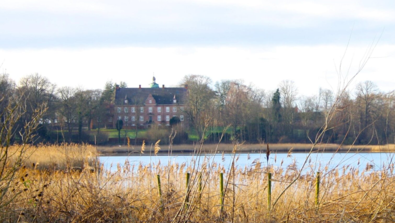 Panoramabillede af en flot sø med en stor rød herregård i baggrunden.