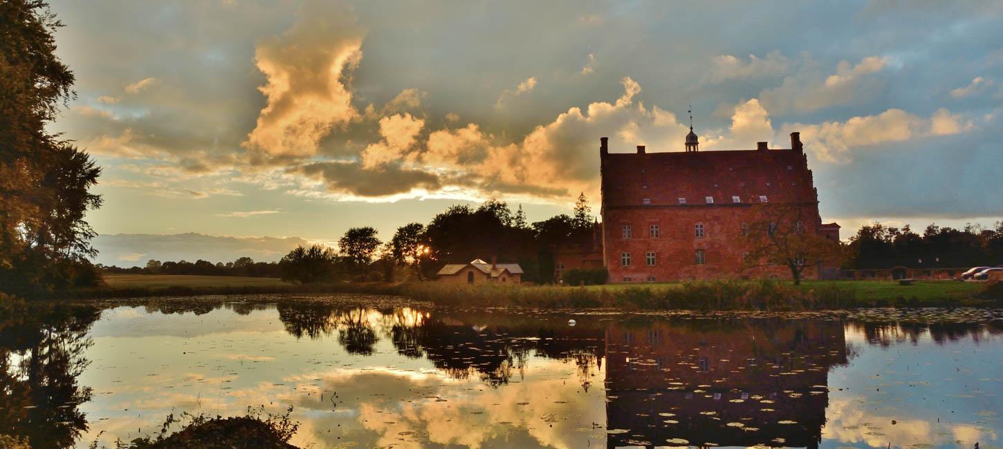 Spejling af det smukke røde slotshotel Broholm Slot i sø med oplyste skyer som baggrund.