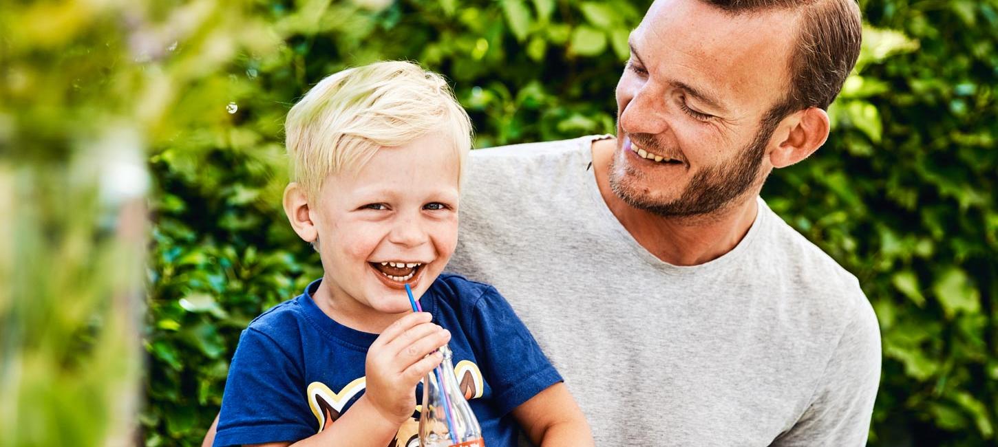 En lille lyshåret dreng sidder på skødet af sin far. Drengen har en blå t-shirt på og en cola med sugerør i hånden. Han smiler stort til kameraet, men hans far kigger smilende ned på ham. De er omgivet er en grøn hæk.