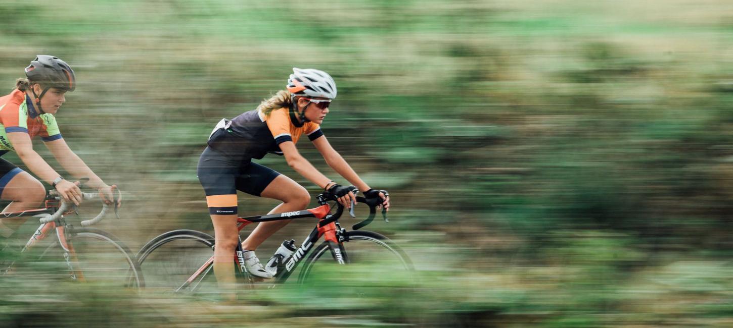 To kvindelige cykelryttere i fuld fart gennem grønt landskab.