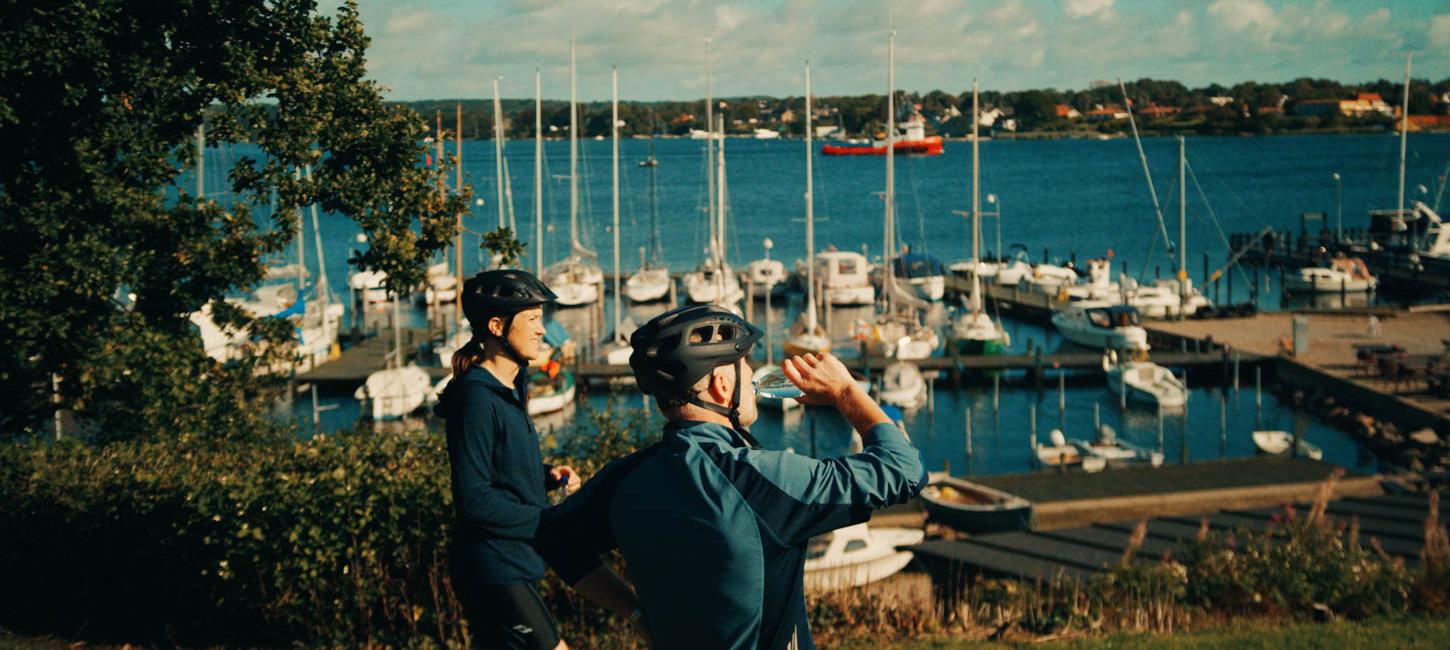 To cyklister med cykelhjelme på får en tår at drikke ved en lille lystbådehavn.