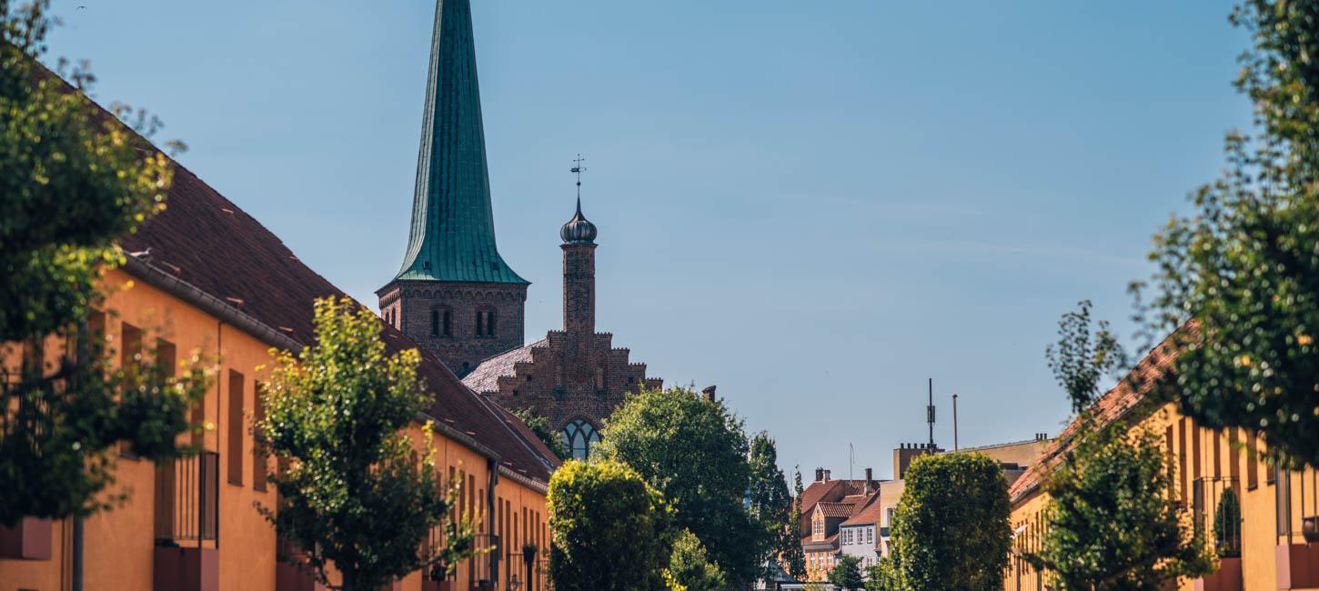 I forgrunden ses toppen af en lang række gule huse med et par træer foran. I baggrunden ses en blå himmel med et kirketårn foran.