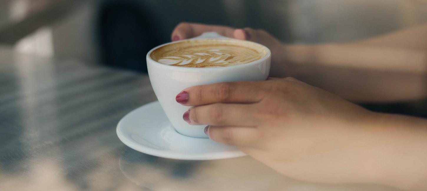 Gennem et vindue er der taget et nærbillede af to hænder der holder en kop kaffe. Hænderne har lyserød neglelak på. Hænderne holder om en hvid kop. I koppen er der kaffe og den er pyntet med latte-art der ligner et blad. Koppen står på en hvid underkop, som står på et lyst træbord.