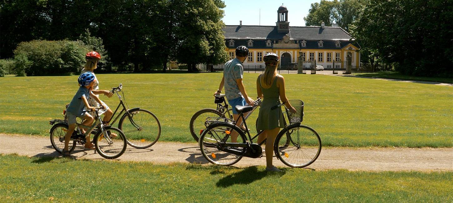 Familie på cykeltur igennem en grøn park, der kigger mod en hvid og gul gård.