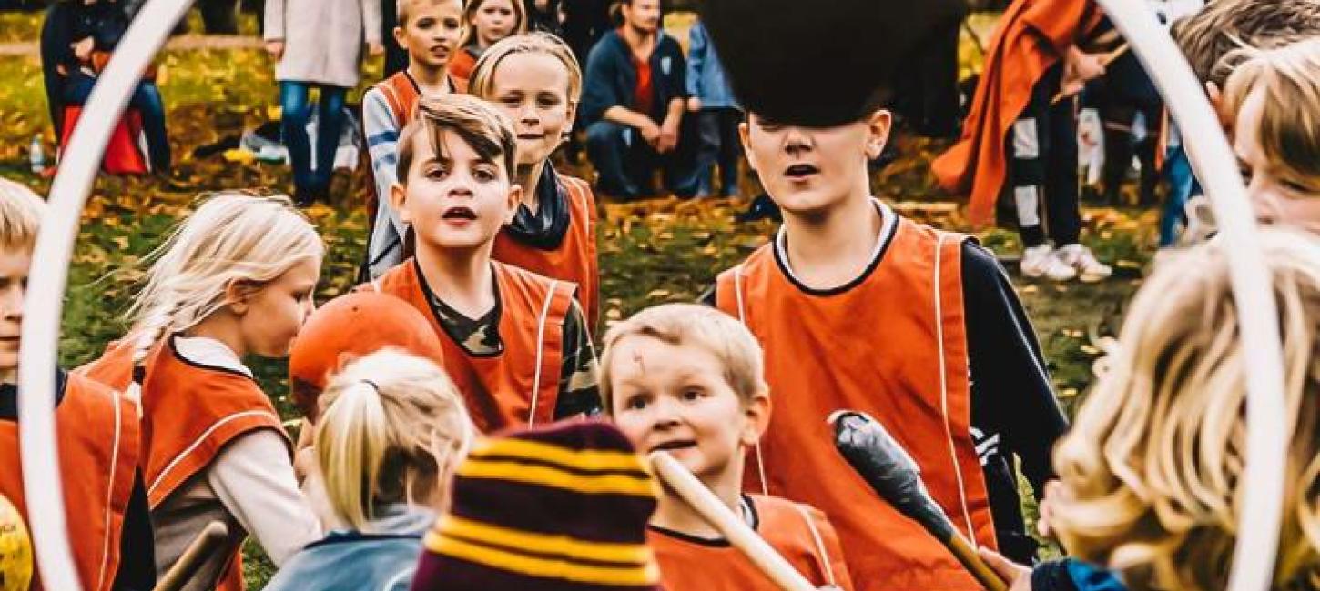 Gennem en hvid hullahopring kan man se en flok børn. Den ene halvdel har røde veste på og den anden blå. De har alle sammen kosteskafter mellem benene og kaster en bold. I baggrunden er forældre tilskuer, mens de står under et kæmpe træ. Børnene spille spillet quidditch fra Harry Potter.