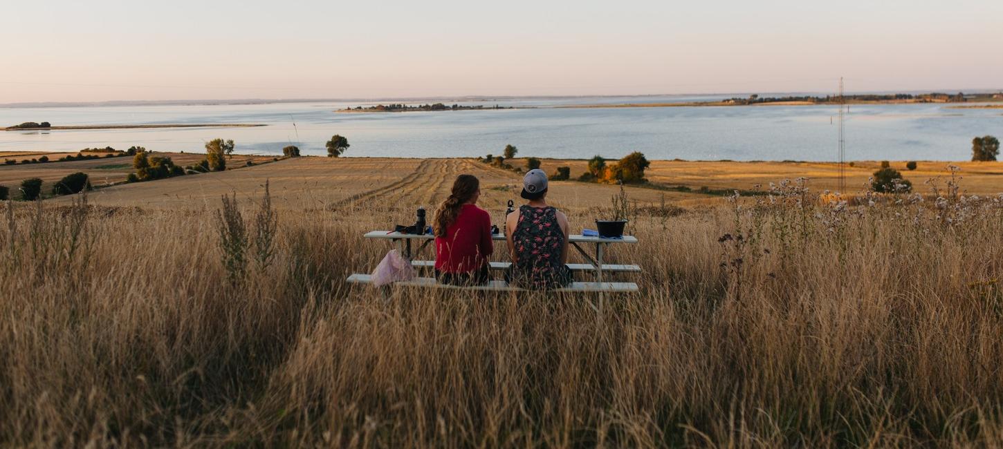 På en mark med udsigt udover havet står et bordebænkesæt. På bænken sidder to kvinder og kigger udover vandet.