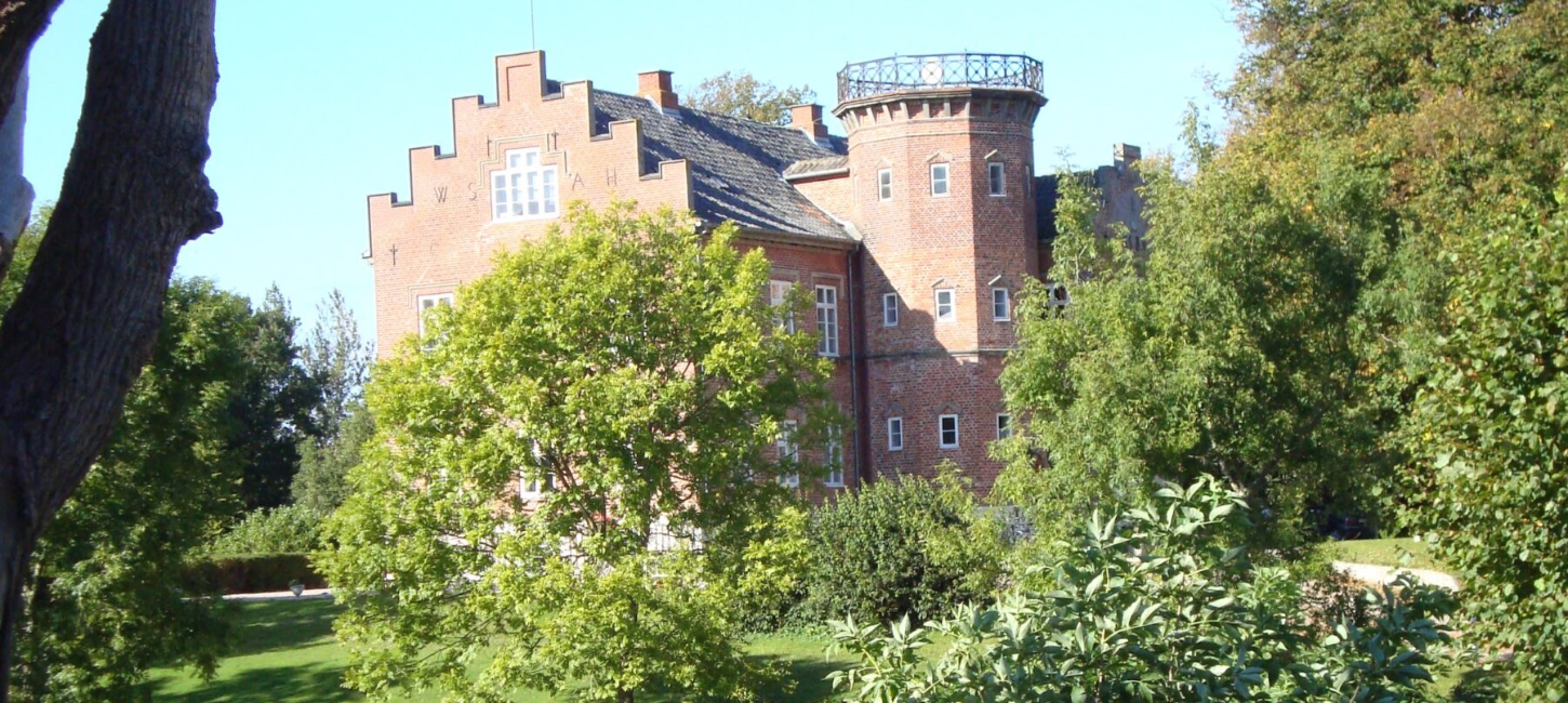 Flot rød herregård med sort tag og et tårn, omkranset af grønt græs og træer.