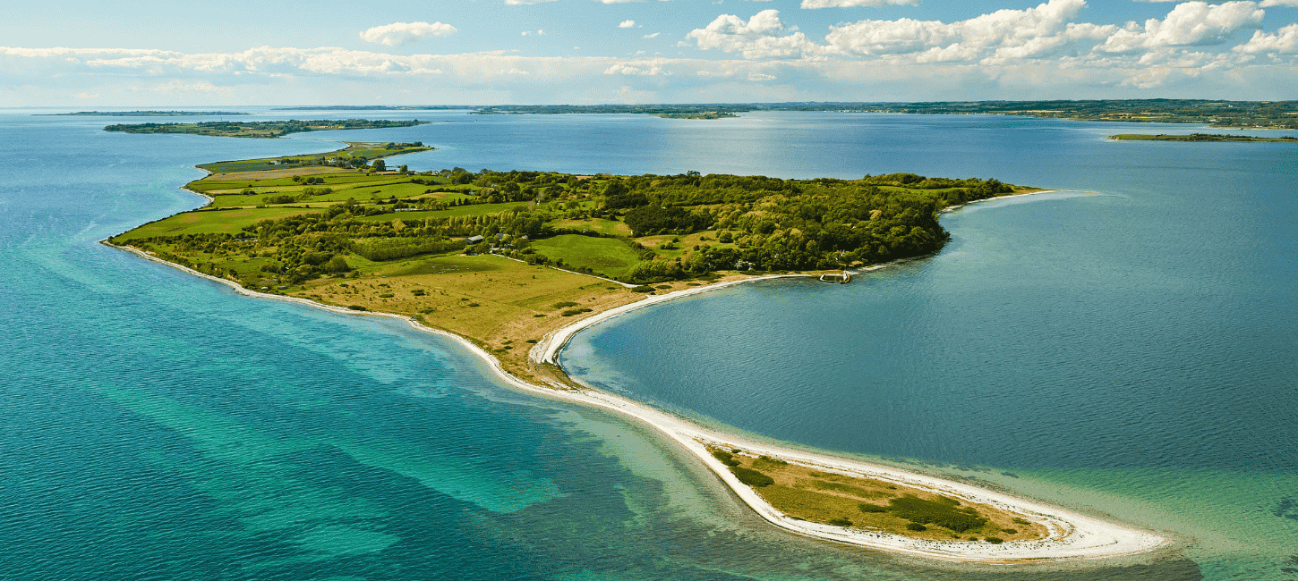 Dronefoto af Avernakø i skarpe, grønne farver med dybblåt vand hele vejen rundt om og horisonten i baggrunden. 