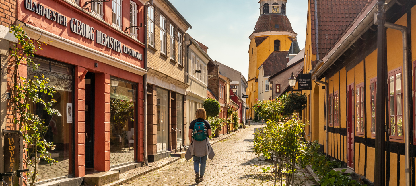 En kvinde med rygsæk går på en smal gade i en by med farverige bindingsværksfacader og et gult klokketårn i baggrunden