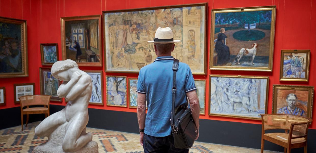 En mand med hat og skuldertaske betragter kunst i et galleri. Han står foran en række malerier på en rød væg, med en skulptur af en nøgen figur til venstre i forgrunden.