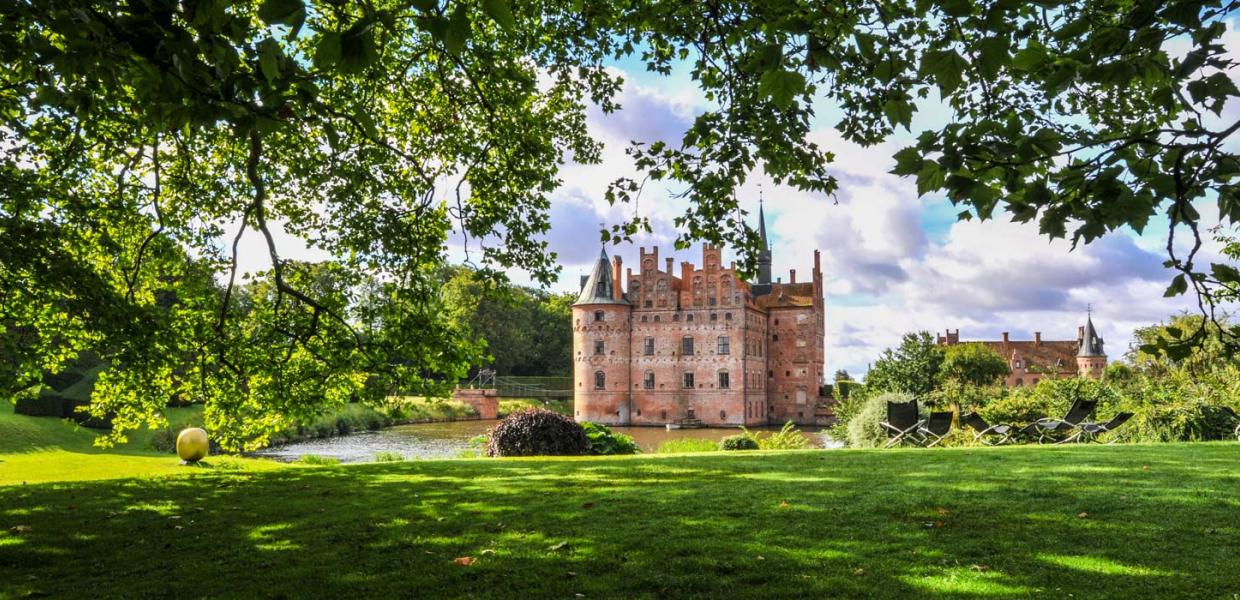 Egeskov slot med grønt græs i forgrunden og grønne træer og blå himmel i baggrunden.