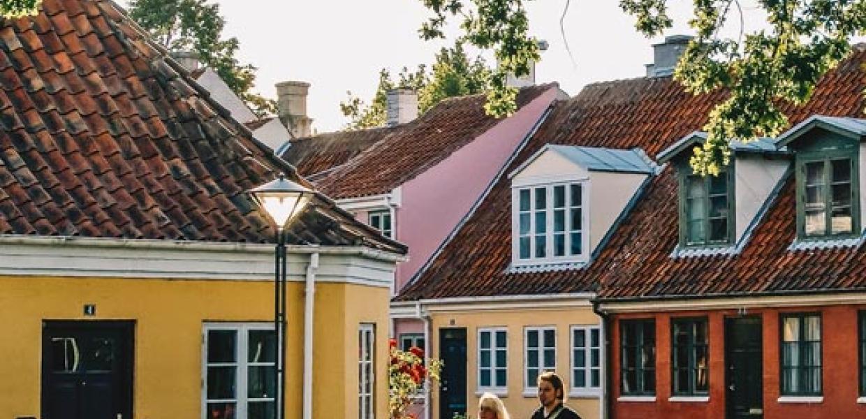 Et par går hånd i hånd på en brostensgade. I baggrundenden er fire bygninger i forskellige farver. Solen skinner gennem en trækrone. 