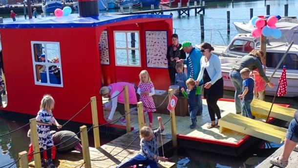En rød åben husbåd er lavet om til en legeplads. Den er fyldt op med legende børn og opmærksomme forældre. I baggrunden ses et par almindelige både tøjret til havnen.
