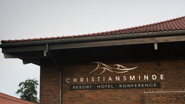 Et nærbillede af et hushjørne hvor logoet til Hotel og konference Christiansminde er i guld. Logoet er tre bølger hvorunder der står Christiansminde, resort, hotel, konference. 