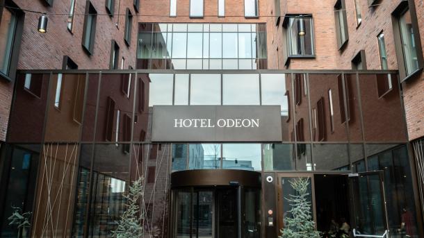Ingangen til Hotel Odeon i Odense. En kobberfarvet svingdør er omringet af store vinduer. Over den hænger et skilt med hotellets logo.