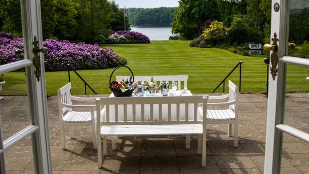 Hindsgavl Slots terrasse med hvide havemøbler. I baggrunden er en lang græsplæne med lilla blomsterbede der fører ned til vandet. 