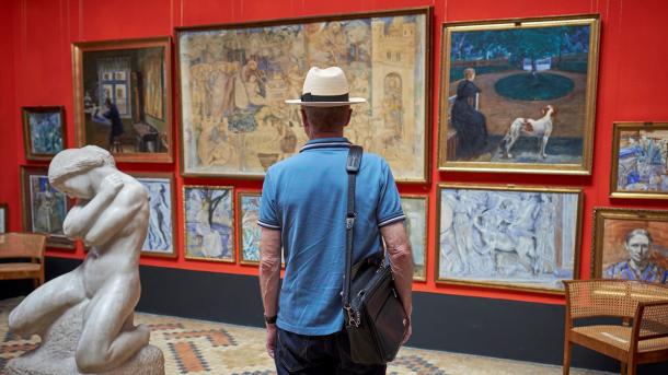 En mand med hat og skuldertaske betragter kunst i et galleri. Han står foran en række malerier på en rød væg, med en skulptur af en nøgen figur til venstre i forgrunden.