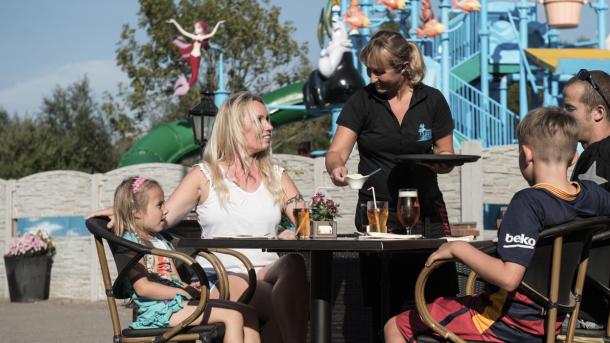 En familie sidder ved et bord, hvor de får drikkevarer serveret. I baggrunden ses vandlandet på Bøsøre strand feriepark