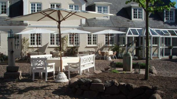 En stor terrasse på en solskinsdag med hvide borde, bænke og parasoller. Bagved er hotel og restaurant Knudsens Gård.