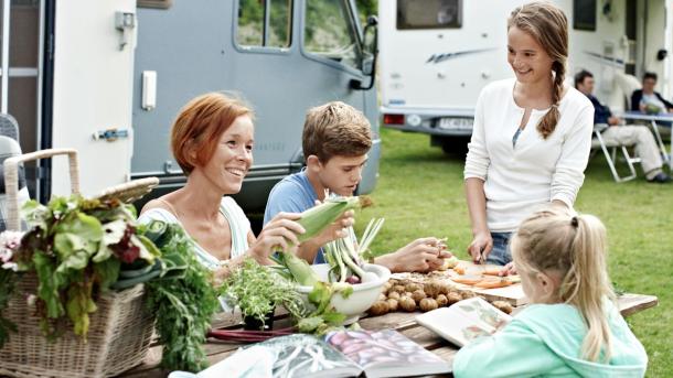 Fire mennesker foran camper sidder og forbereder kartofler, salat og majs.