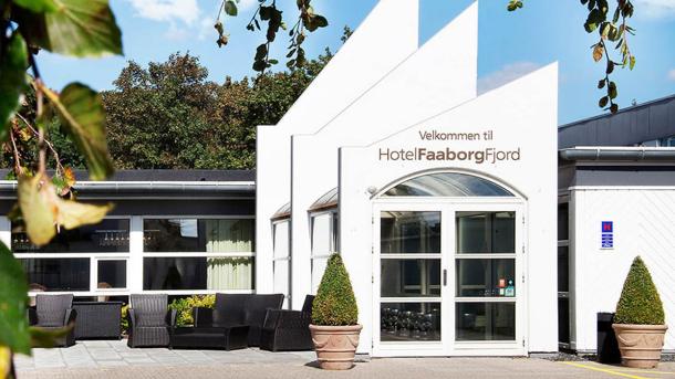 Indgangspartiet til Hotel Faaborg Fjord med en moderne hvid facade og en glasdør. Hotellets navn er fremhævet på bygningens front. Foran hotellet er en terrasse med sorte loungemøbler og to toptrimmede buske i krukker på hver side af indgangen. Billedet er taget på en solrig dag med grønne blade i forgrunden.