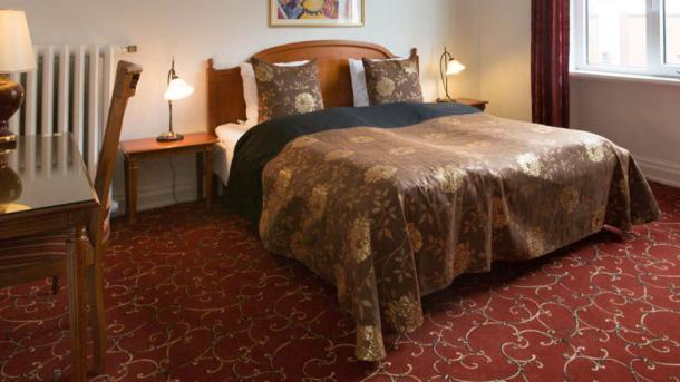En dobbeltseng med brunt sengetæppe og sengegærde i et værelse med rødt mønstret gulvtæppe