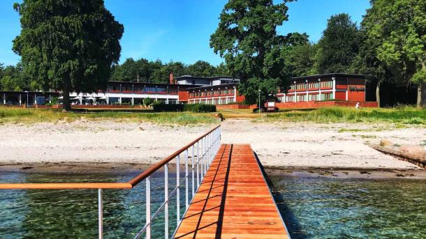 Udefra havet går en træ-badebro ind til sandstranden. I baggrunden ligger det røde Hotel Hesselet gemt bag træerne.