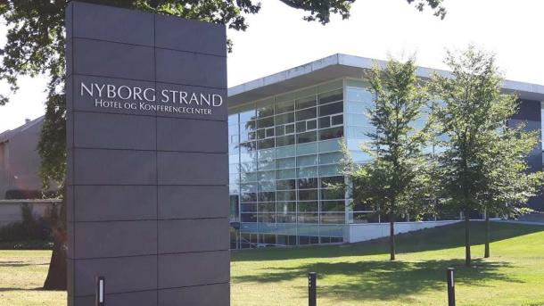 På et højt skilt står der Nyborg Strand Hotel og Konference. Bagved skiltet kan man se en græsplæne med tre træer der står foran en hvid firkantet bygning med mange vinduer.