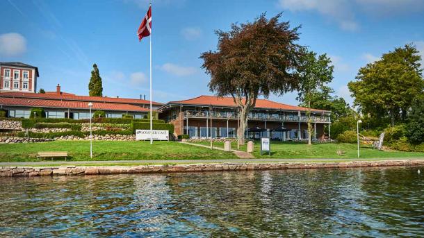 I forgrunden ses havet. I baggrunden er der et rødt hotel. Foran hotellet flagre et dansk flag på flagstangen ved siden af et stort træ.