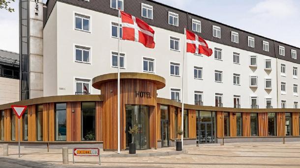 Hvid hotelbygning med sort top. Foran bygningen er et bredt og buet indgangsparti af træ hvor der står Hotel Svendborg. Foran indgangen står to flagstange med det danske flag.