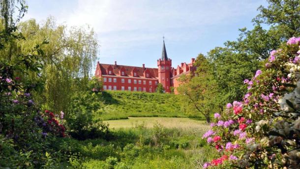 Rødt slot ses på afstand med flot spir og omringet af grønne buske og blomster