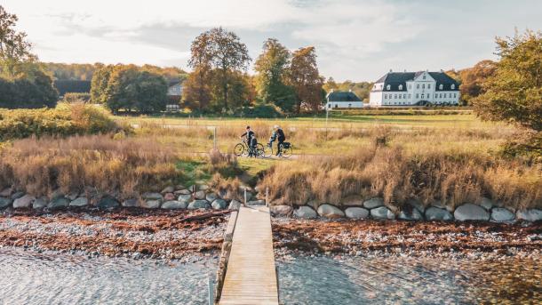 To cyklister gør holdet ved en badebro på strand med dige af store kampesten. I baggrunden en stor, hvid herregård og træer.