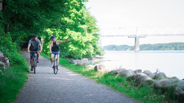 To cyklister kommer kørende ad sti langs med vandet. Den ene peger ud over vandet. En bro anes i baggrunden.