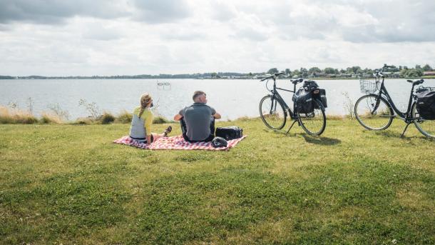 En mand og en kvinder holder rest på et ternet tæppe på græsset med udsigt over vandet. Deres cykler med oppakning står parkeret ved siden af.