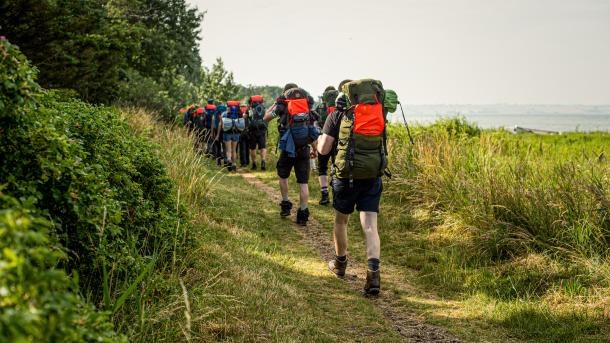 Vandrere med vandrerygsække går på en smal vandresti mellem skov og strand.