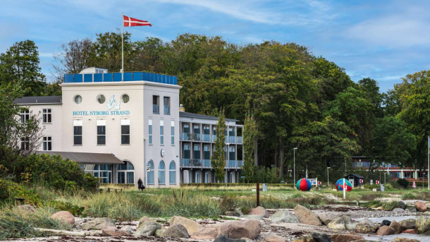 En hvid bygning beliggende tæt ved strandkanten med skov og grønne træer i baggrunden