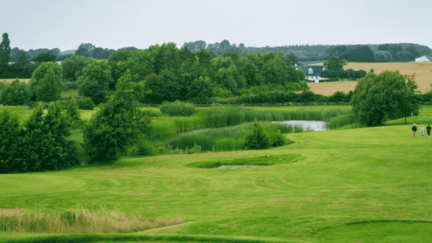 Tre golfspillere går på en bølgende og bakkende golfbane med grønne træer og marker i horisonten