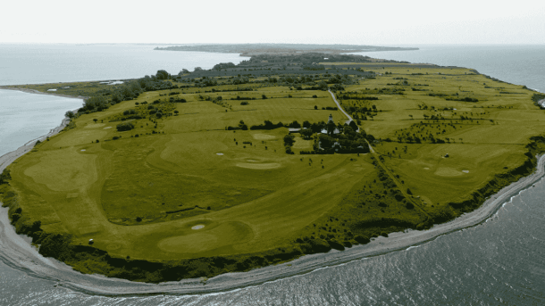 Dronebillede af en ø med en golfbane, der går helt ud til skrænten på en kyst og med et fyrtårn inde i midten