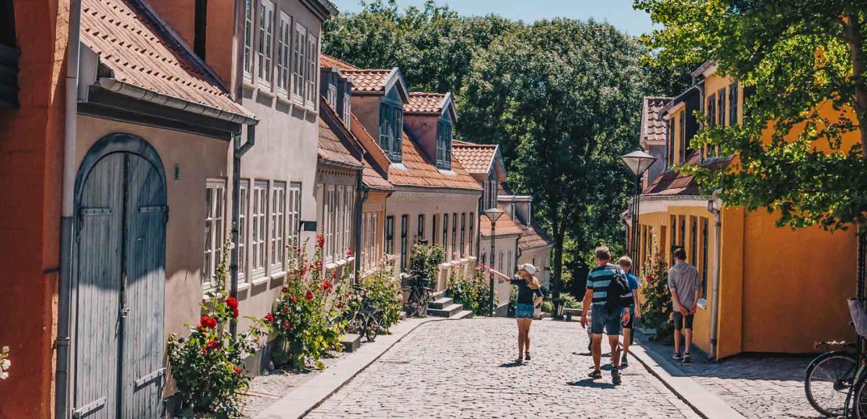 Gammel stræde med brosten og bindingsværkshuse i orange, røde og beige farver. En flok mennesker går tur i det gode vejr og kigger på husene.