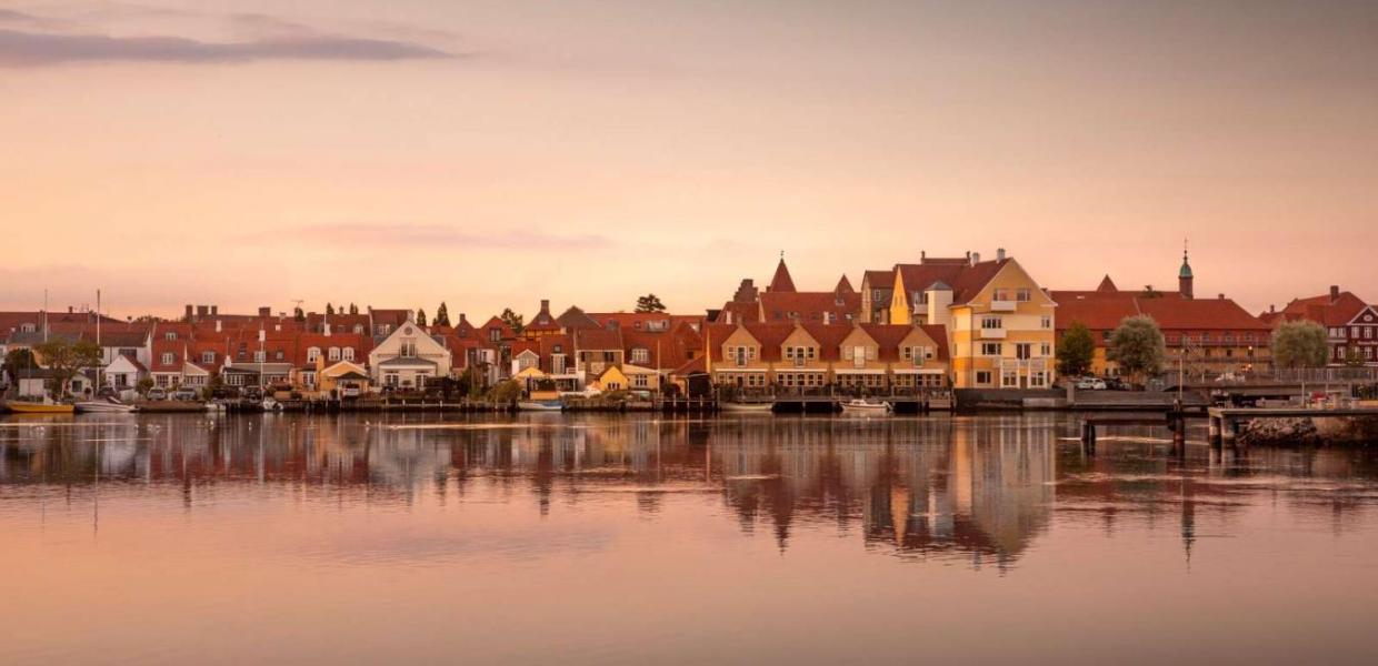 Kerteminde by set fra vandet. Solen er ved at gå nå så himlen er delvist lyserød. I forgrunden er der vand og i bagrunden ses Kerteminde by. Den består af en masse små bygninger i forskellige farver. De har dog alle røde tage.