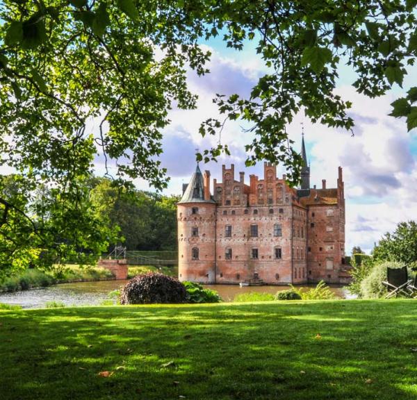 Egeskov slot med grønt græs i forgrunden og grønne træer og blå himmel i baggrunden.